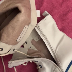 Vita boots med klack från KISSCAT - Säljer ett par vita boots från KISSCAT med kraftig klack och blå detaljer. Skorna har snörning, dragkedja på sidan och dekorativa fyrkantiga nitar. Tillverkade i skinn och har rund tå.