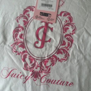 Vit Juicy Couture t-shirt  - Vit t-shirt från Juicy Couture med stort rosa emblem och glittriga detaljer framtill. Klassisk rund halsringning och korta ärmar. Snyggt tryck med JC-logga och dekorativa mönster i rosa. Perfekt för dig som gillar statement-plagg och ikoniska märken.