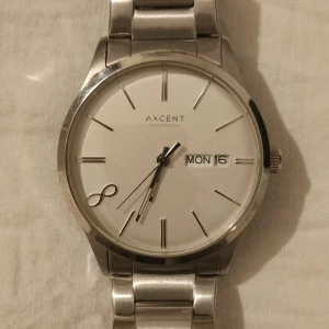 Silverfärgad klocka från Axcent of Scandinavia - Stilren klocka från Axcent of Scandinavia med rund vit urtavla och dag- och datumvisning. Klockan har ett klassiskt armband i silverfärgat rostfritt stål och minimalistiska markeringar på urtavlan.