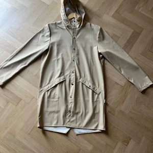 Beige regnjacka från Rains med huva - Helt ny Rains regnjacka har aldrig använt den sedan jag köpt den så. Den är perfekt för dig som vill köpa en stutterheim liknande regnjacka för ett bra pris. Lappar från köp följs med också. Den passar både män och kvinnor då den är unisex som alla regnkappor. Vid funderingar hör av dig✨