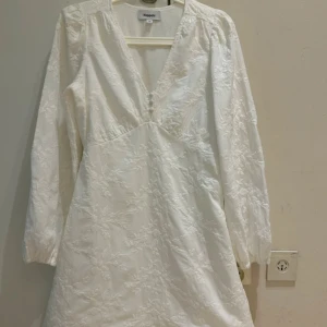 Vit blommig klänning med v-ringning från Kappahl - Säljer en vit klänning från Kappahl i storlek XS. Klänningen har v-ringning, långa ärmar och fina blommiga broderier i tyget. Den har knappar framtill och en lätt utställd passform. Perfekt för sommarens tillfällen! Använd en enda gång 