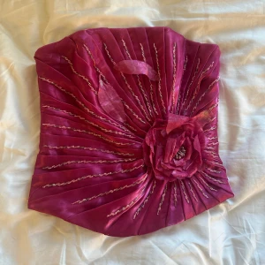 Rosa tubtopp med blomdetalj - Säljer en unik rosa tubtopp med glittriga broderade linjer och en stor tygblomma framtill. Toppen har en glansig finish och är perfekt för dig som vill sticka ut. Snygg till både jeans och kjol!