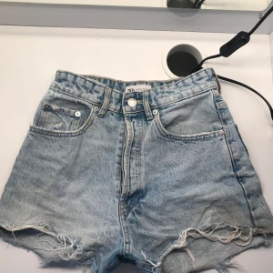 Högmidjade jeansshorts från Zara - Snygga ljusblå jeansshorts från Zara med hög midja och råa, fransiga benslut. Klassisk femficksmodell med dragkedja och knapp framtill. Perfekta för sommaren och ger en avslappnad vibe till din outfit.