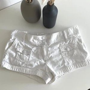 Vita jeansshorts med fickor - Snygga vita jeansshorts med dubbla knappar framtill och fickor med lock både fram och bak. Klassisk design med bälteshällor och kort passform, perfekta för varma dagar. Stilrena och lätta att matcha med allt i garderoben.