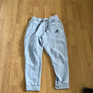 Ljusblå mjukisbyxor från Nike - Säljer ett par ljusblå mjukisbyxor från Nike med svart logga på vänster ben. Byxorna har resår i midjan med snörning och en dragkedjeficka bak. Perfekta för en avslappnad stil. passar perfekt för 146-152