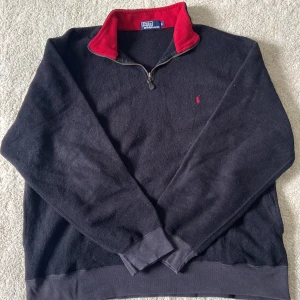 Svart fleece-tröja med dragkedja från Polo Ralph Lauren - Svart fleece-tröja från Polo Ralph Lauren med halv dragkedja och röd insida på kragen. Liten röd logga broderad på bröstet. Perfekt för kyliga dagar och enkel att matcha med andra plagg.