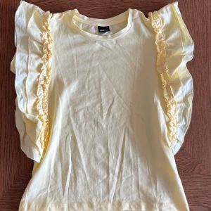 Gul volangtopp från Gina Tricot - Säljer en ljusgul topp från Gina Tricot med korta ärmar och volangdetaljer längs axlarna. Toppen är i mjuk bomull och har en rund halsringning. Perfekt för dig som vill ha något färgglatt och lekfullt i garderoben! Använd endast en gång