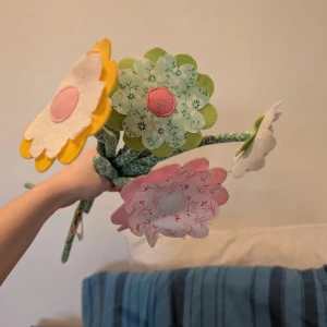 Färgglada tygblommor i bukett - Säljer en bukett med handgjorda tygblommor i olika färger och mönster. Blommorna har mjuka stjälkar och blad i grönt tyg, och blombladen är i rosa, gul, vit, grön och röd med olika mönster som rutigt och blommigt. Perfekt som söt inredningsdetalj!