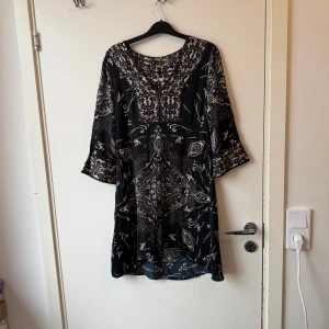Svart mönstrad klänning med trekvartsärm - Säljer en svart klänning med unikt vitt och beige mönster och rundad halsringning. Klänningen har trekvartsärmar och ett lätt, luftigt tyg som faller snyggt. Perfekt för dig som gillar bohemiska detaljer och vill ha något bekvämt men ändå stilrent. Från märket Odd Molly i storlek 0 som enligt deras storleksguide motsvarar ungefär en Storlek XS. Fint skick, men tråddragning finns. 