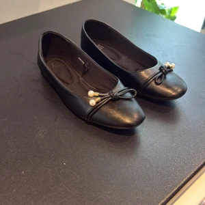 Svarta ballerinaskor med pärldetalj - Stilrena svarta ballerinaskor från XIT med rund tå och dekorativ rosett framtill, prydd med små pärlor och gulddetaljer. Skorna har en platt sula och är tillverkade i syntetmaterial, perfekta för en chic och enkel look.