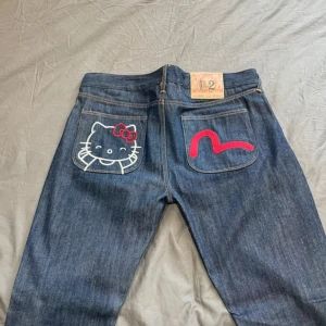 evisu x hello Kitty washed jeans custom - Säljer ett par blå jeans med broderad Hello Kitty på ena bakfickan och röd brodyr på den andra. Klassisk femficksmodell med raka ben och normal passform. Perfekta för dig som vill sticka ut med en lekfull detalj!
