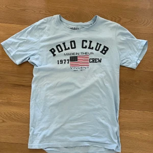 Ljusblå t-shirt från Polo Club - Säljer en ljusblå t-shirt från Polo Club Saint Vincent med tryck framtill och amerikansk flagga. T-shirten har rund hals och korta ärmar. Perfekt för en avslappnad stil. Passar både XS och S