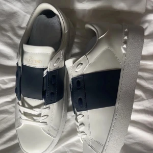 Valentino garavani open - Säljer ett par vita sneakers från Valentino Garavani med bred mörkblå remsa över mitten. Skorna har rund tå, platt vit sula med mönster och klassisk snörning. Märkeslogga på plösen och stilren design. Perfekta för dig som gillar exklusiva sneakers. Köpta här på plick, kvitto finns, säljer pga för små.