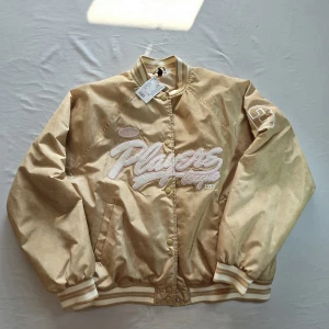 Beige bomberjacka med broderad text från Divided - Säljer en beige bomberjacka från Divided med broderad vit och rosa text 'Players League' framtill. Jackan har långärmad design, ribbade muddar och krage samt tryckknappar. Perfekt för en sportig och trendig look.