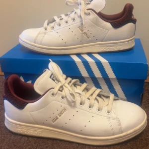 Adidas Stan Smith 'CHENILLE PACK - SHADOW RED' - Säljer ett par klassiska vita Adidas Stan Smith 'CHENILLE PACK - SHADOW RED'. Denna design är väldigt sällsynt och väldigt svåra att få tag på. Skorna har diskret perforering på sidorna och Adidas-logga bak. Perfekta för dig som gillar stilrena och tidlösa sneakers. Hälen är vinröd i ull likt material. 