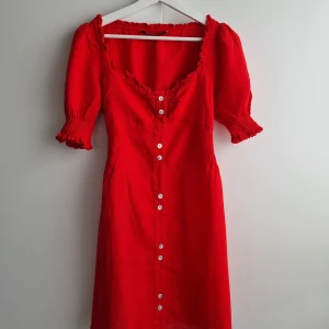 Ny klänning Zara 100% linne - Säljer en röd klänning från Zara i 100% linne. Klänningen har korta puffärmar, fejkknappar framtill och en söt volangkant vid halsringningen. Perfekt för dig som gillar färgstarka och feminina plagg! Helt oanvänd, perfekt skick!