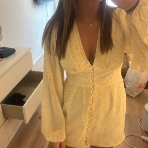 Gul blommig omlottklänning med knappar - Söt kort gul klänning med blommigt mönster, v-ringning och långa puffärmar. Omlottkänsla framtill och dekorativa knappar längs hela framsidan. Perfekt för sommarens alla tillfällen!