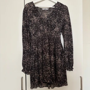 Kortklänning från NA-KD - Snygg kortklänning från NA-KD med unikt mönster i svart och beige. Klänningen har v-ringning, långa ärmar med lätt volang vid ärmslut och smockad midja som ger en fin passform. Perfekt för dig som vill sticka ut med en trendig look.