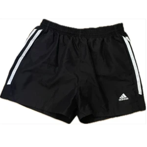 Svarta Adidas träningsshorts - Svarta träningsshorts från Adidas med klassiska vita ränder på sidorna och broderad logga framtill. Shortsen har elastisk midja med snörning. Perfekta för träning på gymmet eller löprundan.