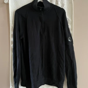 Svart half zip tröja från C.P. Company - Svart långärmad tröja från C.P. Company med ikonisk linsdetalj på ärmen. Tröjan har half zip framtill och hög krage. Tillverkad i mjuk bomull, perfekt för lager-på-lager och streetwear-vibe.