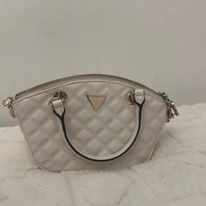 Vit quiltad handväska från Guess Helt ny! - Snygg vit handväska från Guess med quiltat mönster och guldfärgade detaljer. Väskan har två handtag, kedjerem och dragkedja upptill samt en extra ficka med dragkedja på baksidan. Insidan är fodrad med Guess-logga och har flera fack för småsaker. Helt ny väska inte använd. Köpte på Zalando för 800 kr