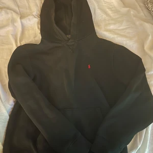 Svart hoodie från Polo Ralph Lauren - Svart hoodie från Polo Ralph Lauren med klassisk röd logga broderad på bröstet. Tröjan har huva och långa ärmar. Perfekt för dig som gillar en clean och stilren look. Materialet är mjuk bomull, TAR BYTEN