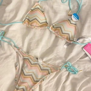 Pastellfärgad zigzag bikini  - Superfin bikini  i pastelliga färger med zigzag-mönster i ljusblått, rosa, guld och grått. Klassisk trekantsmodell med knyt i sidorna och avtagbar vaddering. Helt oanvänd därav priset. Från New yorker i Italien, tror inte den finns i sverige, men inte helt säker, därav priset.