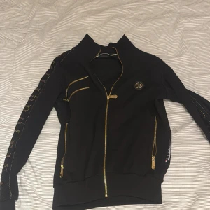 Philipp Plein x Mayweather - Svart hoodie från Philipp Plein x Floyd Mayweather med guldiga dragkedjor och stora glittriga guldtryck på ryggen och ärmarna. Jackan har ståkrage, flera fickor med dragkedja och exklusiva detaljer som PP-logga på bröstet. Perfekt för dig som gillar statement-plagg. Hör av er vid frågor , pris kan diskuteras 