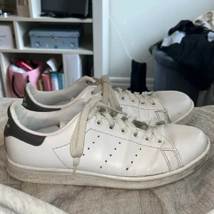 Adidas Stan Smith vita sneakers - Klassiska Adidas Stan Smith sneakers i vitt skinn med svarta detaljer på hälen och perforerade ränder på sidorna. Skorna har rund tå, platt sula och snörning framtill. Perfekt för en clean och tidlös look.