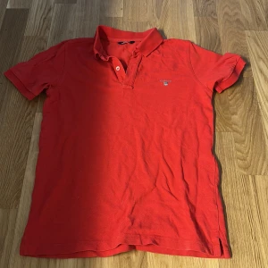 Röd pikétröja från Gant - Klassisk röd pikétröja från Gant med korta ärmar och krage. Tröjan har två knappar vid halsen och en liten broderad Gant-logga på bröstet. Perfekt för en avslappnad men ändå stilren look.