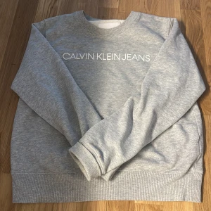 Grå sweatshirt från Calvin Klein Jeans - Snygg grå sweatshirt från Calvin Klein Jeans med vit logga tryckt över bröstet. Klassisk rund halsringning och långa ärmar med ribbade muddar. Perfekt för en chill och stilren look.