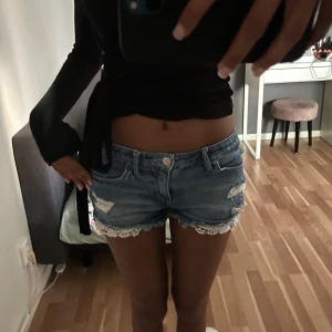 Blå jeansshorts med spetskant - Snygga blå jeansshorts med slitna detaljer och vit spetskant längst ner. Klassisk femficksmodell med knapp och dragkedja framtill. Perfekta för sommaren och ger en cool, avslappnad vibe.