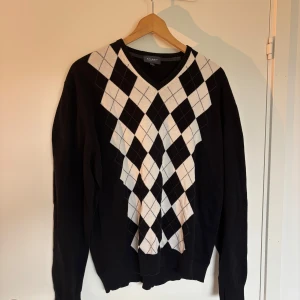 Svart och vit rutig tröja från Atlant - Snygg svart tröja från Atlant med klassiskt rutig argyle-mönster i vitt och svart framtill. V-ringad halsringning och långa ärmar. Tillverkad i mjuk bomull som känns skön mot huden. Perfekt för dig som gillar preppy stil och vill sticka ut lite extra.