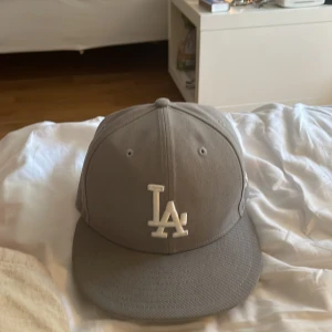 Grå LA-keps från New Era 59FIFTY - Snygg grå keps från New Era 59FIFTY med LA-logga broderad framtill i vitt. Klassisk basebollmodell med böjd skärm och broderade ventilationshål. Kepsen är i bomull och har MLB-logga baktill. Perfekt för dig som gillar streetwear och sportig stil 56 cm runt huvudet .