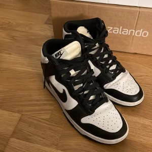 Nike Dunk High svartvita sneakers - Svartvita Nike Dunk High sneakers med klassisk siluett och högt skaft. Skorna har svart swoosh, svarta snören och perforerad tåbox för extra ventilation. Tillverkade i skinn med vit bas och svarta paneler. Perfekta för dig som gillar streetstyle och vill sticka ut.