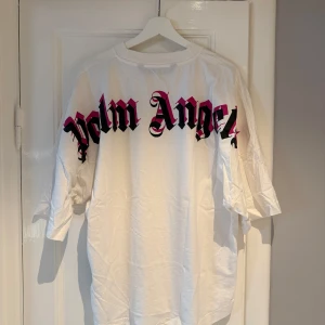 Vit oversized Palm Angels t-shirt - Vit oversized t-shirt från Palm Angels med stort svart och rosa logotryck över ryggen och mindre logga i nacken. T-shirten har en loose passform, rund hals och är tillverkad i mjuk bomull. Perfekt för en streetwear-look. (Shoutout AI) M/L