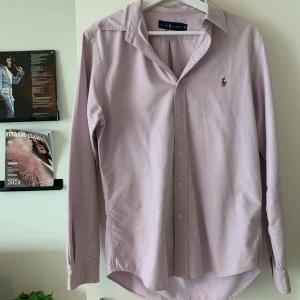 Ljuslila skjorta från Ralph Lauren - Snygg ljuslila skjorta från Ralph Lauren i slim fit-modell. Klassisk krage, knäppning framtill och broderad logga på bröstet. Skjortan är långärmad och tillverkad i bomull, perfekt för en clean och stilren look.