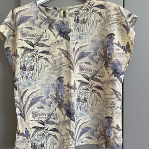 Beige blus  - Unik beige blus . Mönstret har tropiska växter och människor, samt textinslag. Blusen har rund halsringning och knapp bak i nacken. Tillverkad i en mjuk viskosblandning.