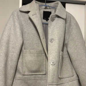 Ljusgrå kappa med stora fickor - Stilren ljusgrå kappa med klassisk krage och stora, fyrkantiga fickor framtill. Kappan har knäppning med tre stora knappar och är helfodrad. Perfekt för dig som gillar en clean och enkel look.