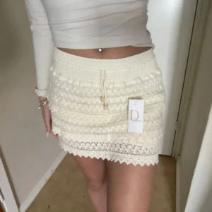 Vit stickad minikjol med snörning - Supersöt vit stickad minikjol från Daphne med volangdetaljer och snörning i midjan. Kjolen har virkade mönster och en elastisk midja som gör den bekväm och lätt att styla. Perfekt för en fräsch och somrig look!