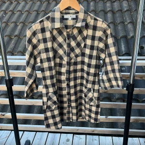 Rutig skjorta från Think Positive M - Snygg rutig skjorta från Think Positive i svart och beige. Skjortan har trekvartsärm, klassisk krage, knappar framtill och två fickor. Tillverkad i ett lätt bomullstyg som känns skönt mot huden. Perfekt för dig som gillar en avslappnad men trendig stil.