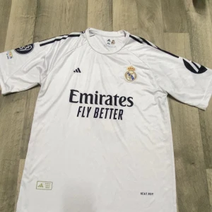 Real Madrid Mbappé 9 matchtröja - Säljer en vit Real Madrid fotbollströja från Adidas med Mbappé och nummer 9 på ryggen. Tröjan har korta ärmar, klubbmärke på bröstet och sponsortryck framtill. Tillverkad i lätt och ventilerande funktionsmaterial, perfekt för match eller träning.