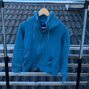 Blå zip-hoodie från AC, XS - Säljer en blå hoodiejacka från AC i storlek XS. Jackan har dragkedja, hög krage med snörning och broderad text 'UNITED' på baksidan av kragen. Två fickor framtill med dragkedja. Perfekt för dig som gillar en chill och sportig stil.