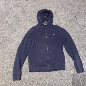 Marinblå hoodie från Lyle & Scott - Marinblå hoodie från Lyle & Scott med dragkedja, huva med snörning och klassisk gul broderad logga på bröstet. Tröjan har känguruficka framtill och ribbade muddar vid ärmslut och nederkant. Perfekt för en chill och stilren look.