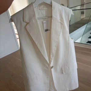 Vit kostymväst från H&M, XS - Stilren vit kostymväst från H&M i storlek XS. Västen har klassisk krage, en knapp framtill och två fickor med lock. Perfekt för att lyfta din outfit med en clean och modern look. Materialet är mjukt och känns lyxigt mot huden.