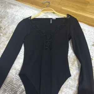 Svart ribbad body med spets från H&M - Svart långärmad body från H&M Divided i ribbat material. Djup v-ringning med dekorativ spetsdetalj längs halsringningen och framtill. Storlek xs
