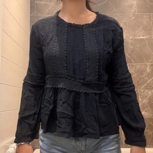 Svart blus med spetsdetaljer - Supersöt svart blus med volang nertill och spetsdetaljer längs ärmar och framtill. Långärmad modell med lätt puff och struktur i tyget. Perfekt till jeans för en chill men ändå uppklädd look.