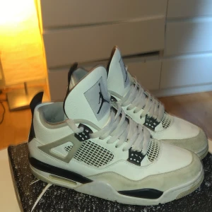 Nike Air Jordan 4 Retro Vit/Svart - Säljer ett par Nike Air Jordan 4 Retro i vitt och svart med grå detaljer. Skorna har klassisk meshpanel, snörning och ikonisk Jumpman-logga på plös och häl. Ovandelen är i skinn och mocka, med robust sula och coola plastdetaljer på sidorna.