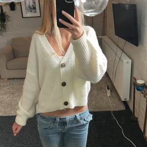 Vit stickad kofta från Vero Moda - Superfin vit stickad kofta från Vero Moda med stora bruna knappar och v-ringad hals. Modellen är croppad och har långärmade ärmar med breda muddar. Perfekt att slänga över ett linne för en chill och trendig look.