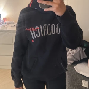 Svart hoodie från Hoodrich med tryck - Svart hoodie från Hoodrich med stor grå logga framtill och coola tryck på huvan och ryggen. Hoodien har huva med röda detaljer, känguruficka och långa ärmar. Perfekt för dig som gillar streetwear och vill sticka ut lite extra.
